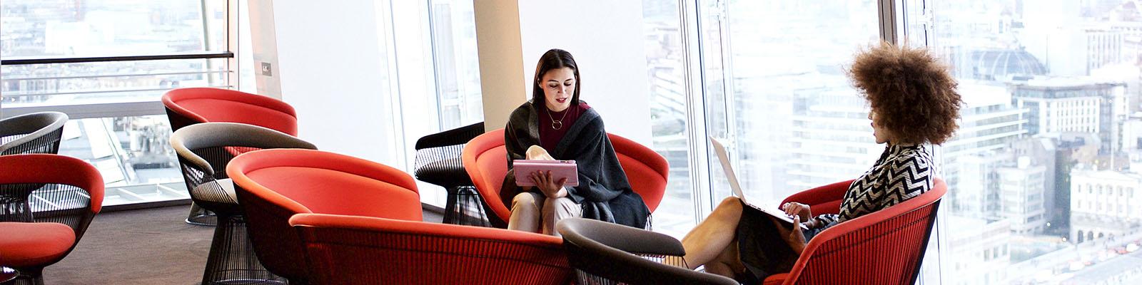 Photo of two women seated in red chairs, holding digital devices on their laps as they talk.