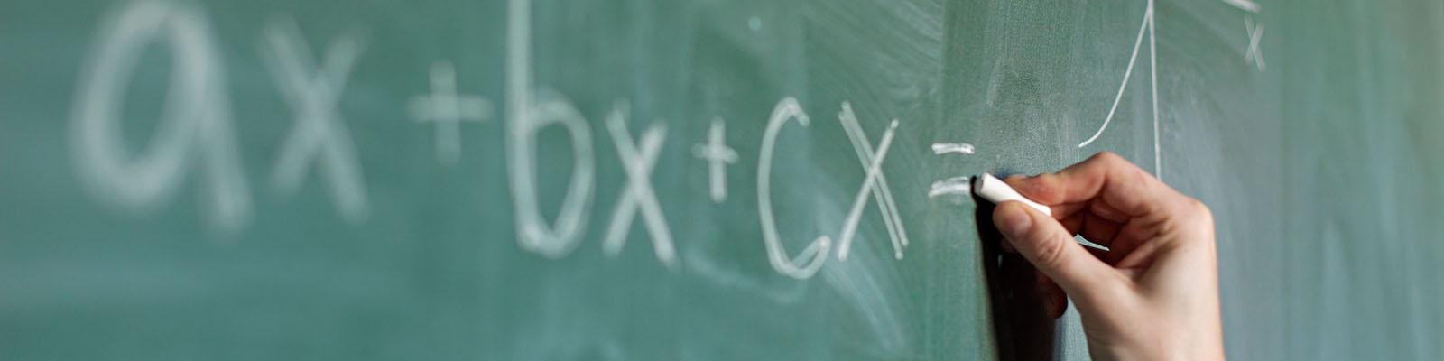 Image of a green chalkboard, with a hand writing mathematical equations in chalk. 